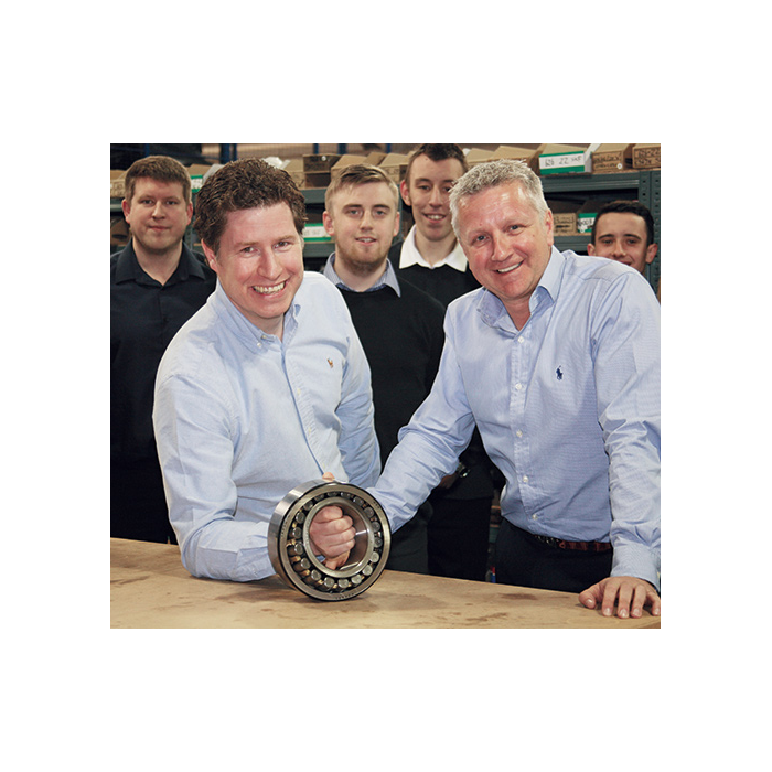 Quality Bearings Online Limited Directors Denny and Simon holding a bearing in the warehouse with four employees behind them.